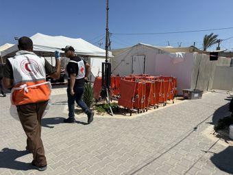 The photo shows two people wearing Red Cross vests walking along a paved path at a field hospital, with white tents and equipment visible around them. Several large red containers are lined up outside the tents under a clear blue sky.