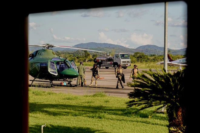 The image shows a group of people in military or police uniforms at an airport. They are standing next to a green helicopter, and there is also a car and a small aircraft in the background. The surrounding landscape features green grass, palm trees, and distant mountains beneath a blue sky with a few clouds