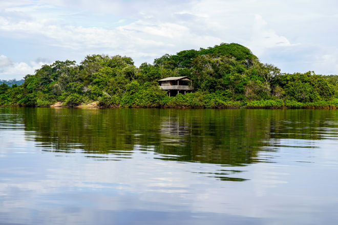 The image shows a lush, green island with dense vegetation. In the middle of the island stands a simple, wooden house on stilts, partially hidden by the trees. The island is surrounded by calm water that reflects both the house and the greenery. The sky is bright with a few clouds, and the landscape evokes a sense of tranquility and isolation, far from any city or town.