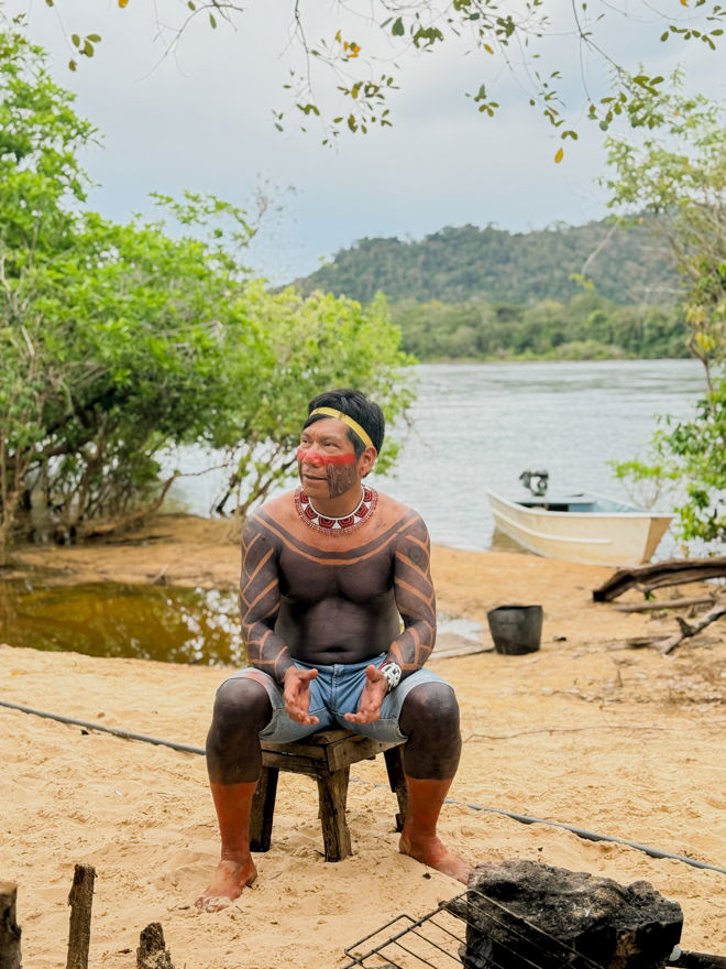 A man is sitting on a simple wooden chair on a sandy riverbank. His upper body, arms, and legs are covered in extensive black and red body paint patterns.
