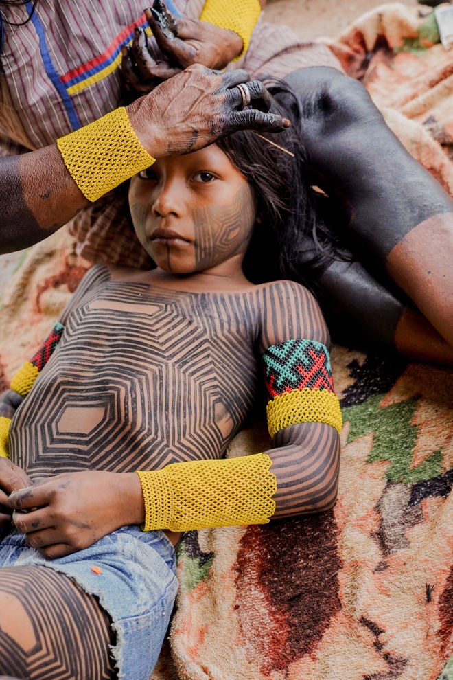 The image shows a child lying on a blanket, while an adult woman sits beside them, tending to the child's hair. The child has geometric black body paintings covering the torso, arms, and legs, and is wearing denim shorts.