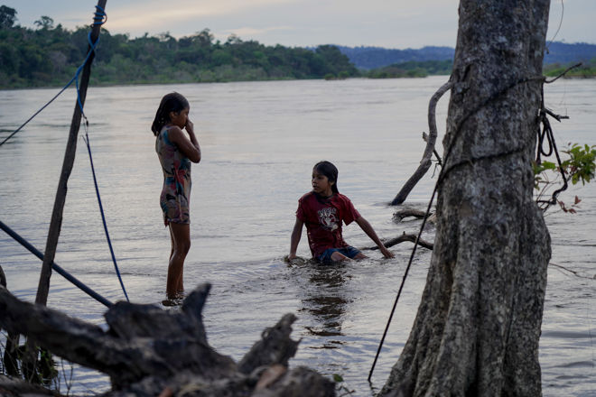 The image shows two children by the riverbank in a natural landscape. One is standing in the water wearing a short summer dress, while the other is squatting in the water wearing a red T-shirt and shorts. The water is calm, and there are trees and vegetation around them, with a forest in the background