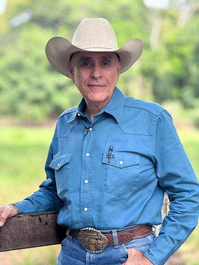 An elderly man wearing a cowboy hat and a blue shirt is standing by a wooden fence with green trees in the background.