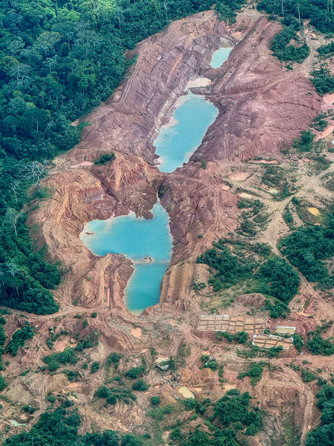 The image shows a large area of deforested and excavated land, likely an open-pit or gold mine in the rainforest. In the middle of the site are several large blue-green ponds surrounded by red and brown soil, with remnants of forest along the edges