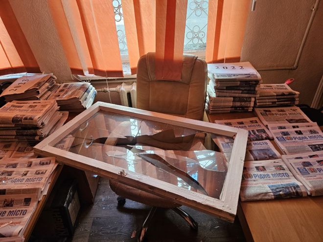 The image shows an office space with stacks of newspapers piled on desks and tables. Some of the bundles are labeled with years such as "2022" and "2023." In the center, there is a large chair and a broken window frame with shattered glass resting on it. The background features orange vertical blinds, and the overall setting looks cluttered and somewhat chaotic.