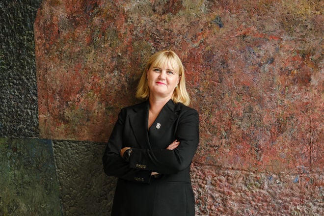 A woman with light, shoulder-length hair stands in front of a colourful wall, wearing a dark blazer with a brooch.