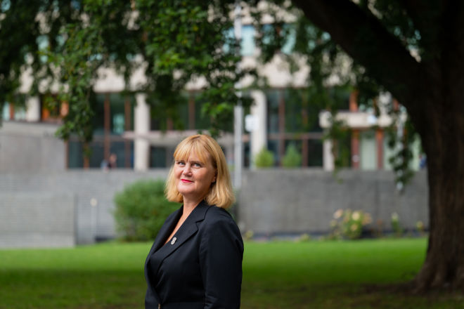 A woman with blonde hair and a dark blazer is standing on a lawn in front of a building and a large tree.