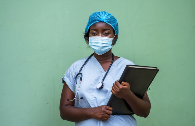 Nurse in blue scrubs with a stethoscope around the neck holding a black binder, standing against a green wall.