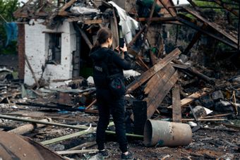 A woman in dark clothing stands among debris and damaged buildings, taking a photo with her phone.