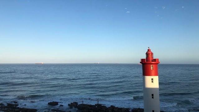 og denne  Bildet viser en vakker kystscene med en rød og hvit fyrlykt som står til høyre i forgrunnen. Fyret er slankt og høyt, og det skinner i sollyset. Bakgrunnen består av en uendelig havflate som strekker seg mot horisonten, hvor det er noen få skip som kan sees langt i det fjerne. 