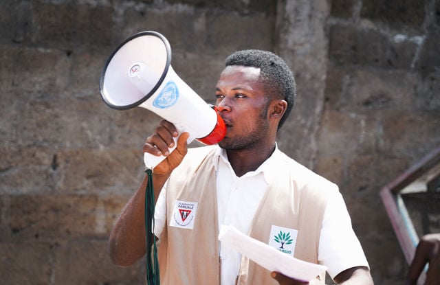 An adult man holds a megaphone with a UN logo in front of a stone wall.