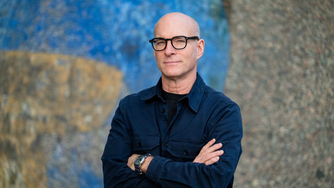 A bald man with glasses stands with arms crossed, wearing a dark jacket in front of a textured, colorful wall.