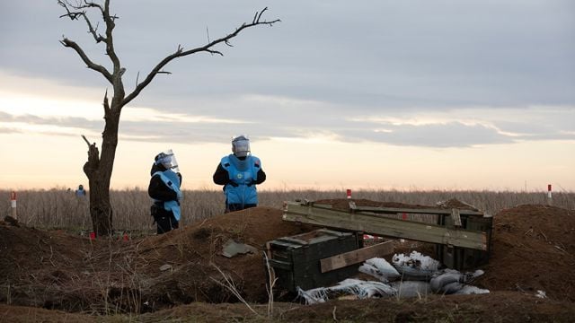 Two deminers wearing protective gear stand in a field at sunset, surrounded by marked posts indicating a dangerous area. A leafless tree stands nearby, and the landscape is open and flat under a cloudy sky.