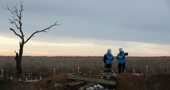 This image depicts a bleak, open landscape under a cloudy sky, with the sun setting or rising on the horizon. In the foreground, there is a single, leafless tree with twisted branches, adding a sense of desolation to the scene. Two people stand nearby, both wearing protective blue vests and helmets that cover their entire heads, suggesting they are involved in dangerous work—possibly demining or bomb disposal. 