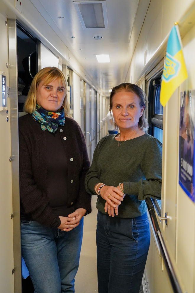 Two adult women are standing in the corridor of a train next to a window with a small Ukrainian flag.