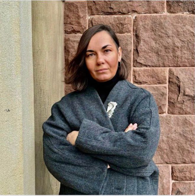 A woman with shoulder-length brown hair stands with arms crossed, wearing a dark coat in front of a brick wall.