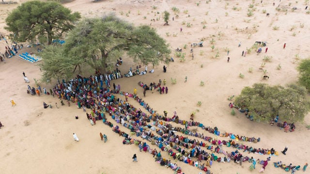 The image depicts a large group of people sitting in long, parallel rows on a dry, sandy open plain. There are a few scattered trees providing some shade. The people appear to be gathered for an event or activity, and most are dressed in colorful clothing.