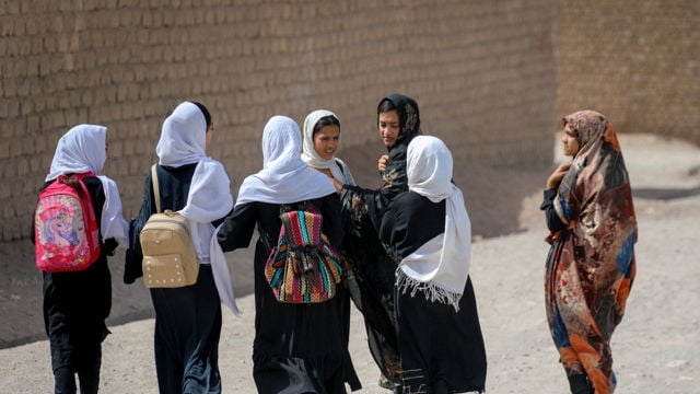 The image shows a group of young girls walking together along a road. They are dressed in traditional clothing, including long dresses and headscarves. Some of the girls are carrying colorful backpacks. 
