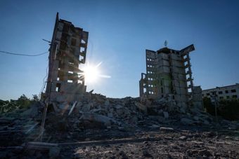 The photo shows two destroyed high-rise buildings standing among large amounts of rubble and debris. The sun shines low between the damaged structures, and the sky is clear and blue. The atmosphere is marked by silence and devastation, suggesting the area has suffered extensive damage.