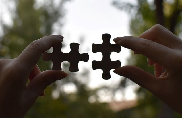 The image shows two hands, each holding a puzzle piece. The hands are facing each other, and the pieces are held in a way that they almost fit together, but are still slightly apart.
