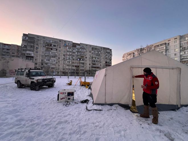 A person in a red jacket stands outside a large tent in a snowy area, with an off-road vehicle and apartment buildings in the background.