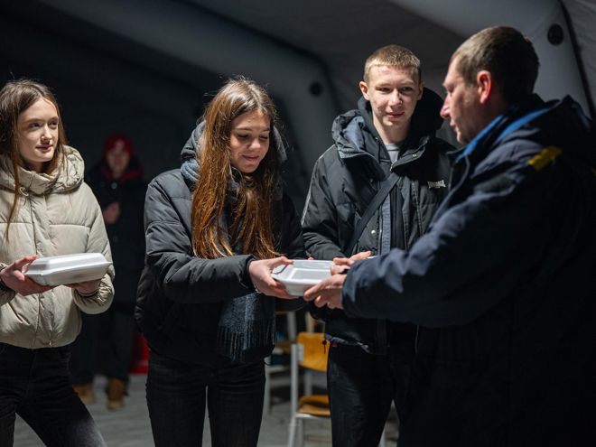 Several young people in jackets receive food containers from a person in uniform inside a tent.    Copy response