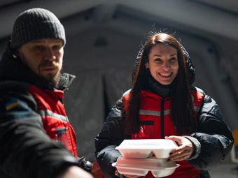Two adults wearing red jackets are handing out food in takeaway containers indoors.
