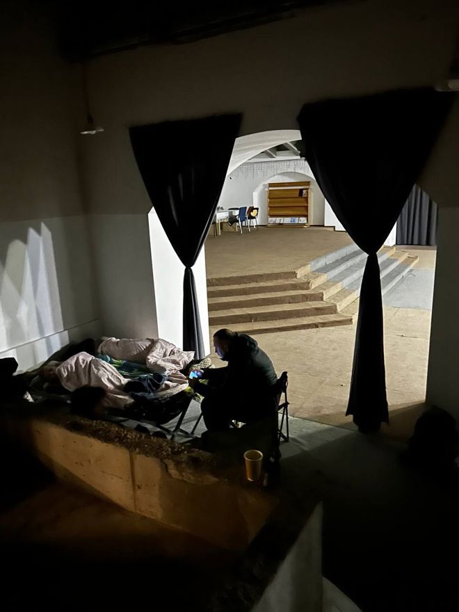 A person sitting with a phone while two other people are sleeping inside of a church building. 