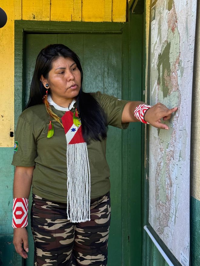 A woman stands in front of a wall, pointing at a large map that is hanging up. She has long, dark hair and is wearing a green T-shirt with a Brazilian flag on the sleeve, camouflage-patterned pants, and colorful red and white bracelets.